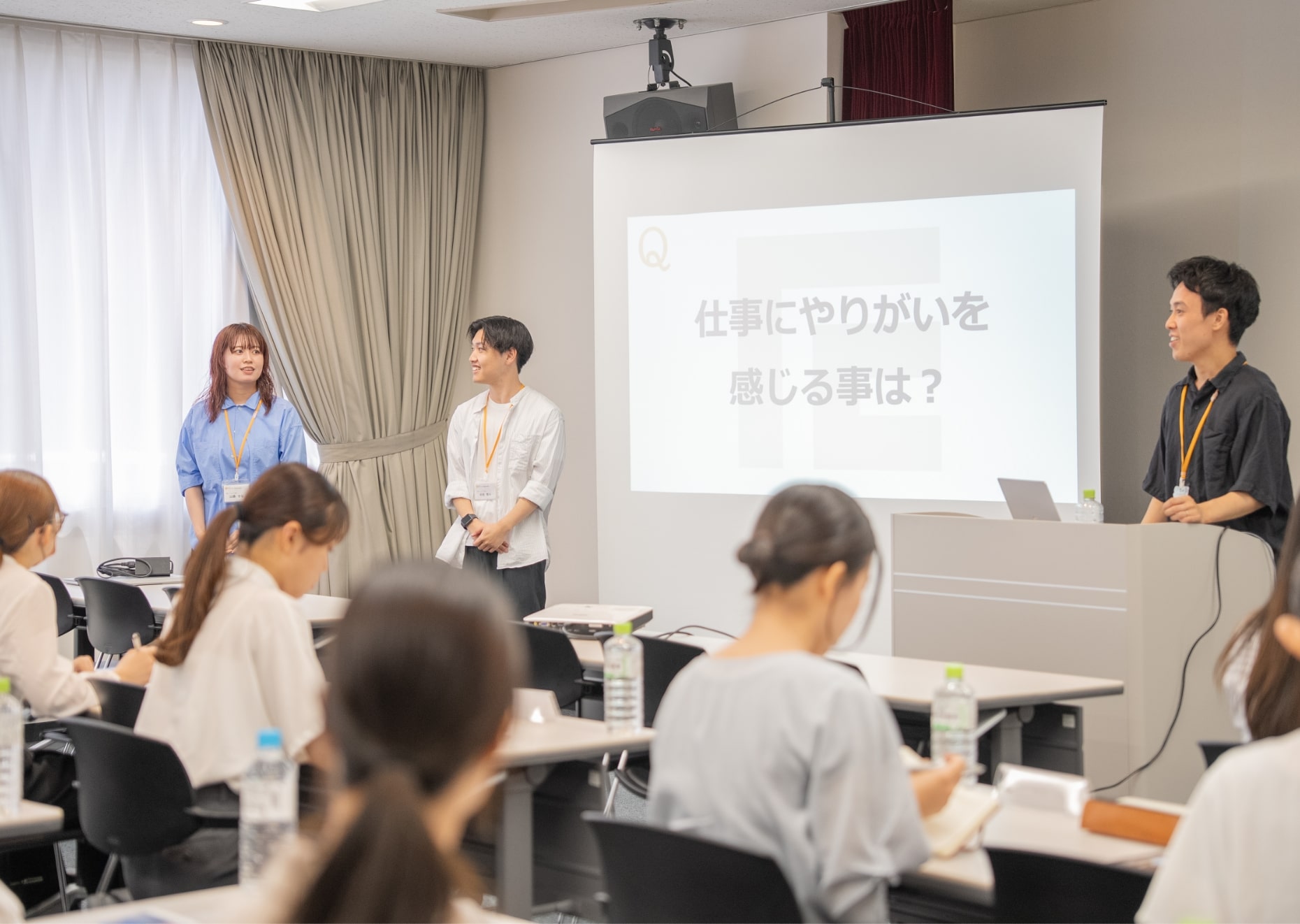 数人の参加者が椅子に座り、発表者3人がプレゼンを行っている場面